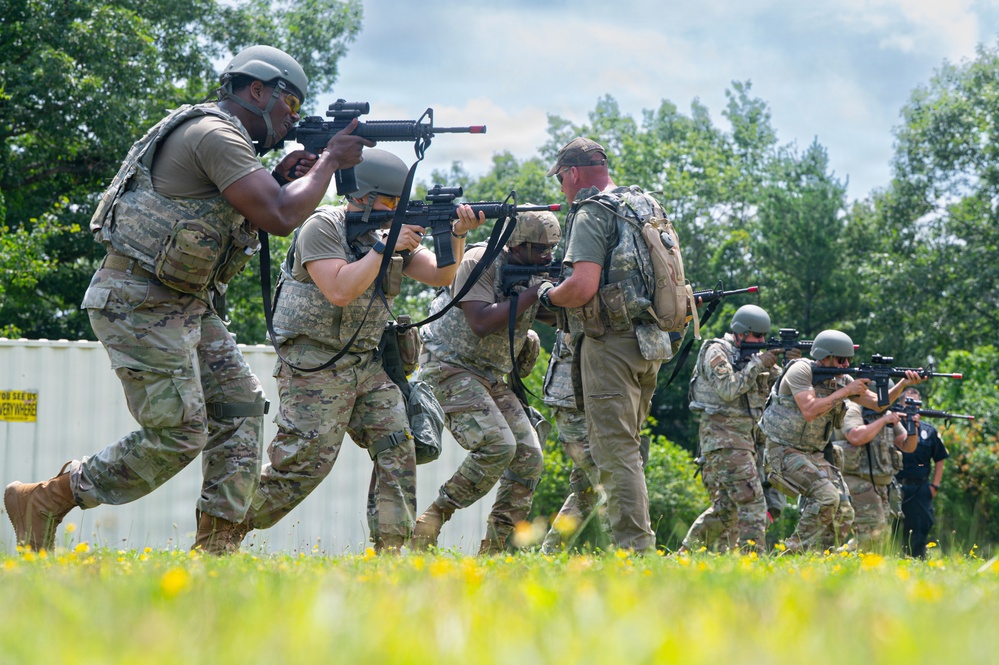 A group of military personnel in camouflage uniforms are conducting a tactical training exercise in an outdoor field setting. The soldiers are moving in formation while aiming rifles, suggesting a simulated assault or clearing drill. The setting appears to be a designated training area with a white structure and natural vegetation in the background.