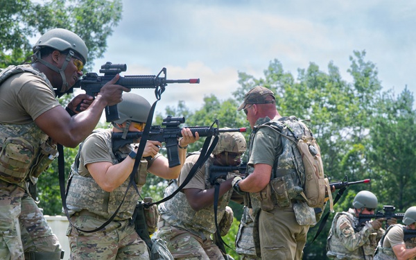 A group of military personnel in camouflage uniforms are conducting a tactical training exercise in an outdoor field setting. The soldiers are moving in formation while aiming rifles, suggesting a simulated assault or clearing drill. The setting appears to be a designated training area with a white structure and natural vegetation in the background.