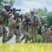 A group of military personnel in camouflage uniforms are conducting a tactical training exercise in an outdoor field setting. The soldiers are moving in formation while aiming rifles, suggesting a simulated assault or clearing drill. The setting appears to be a designated training area with a white structure and natural vegetation in the background.