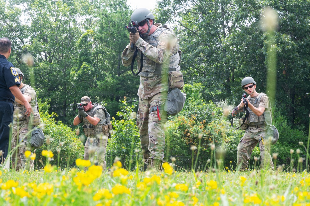 The image shows a group of tactical personnel in military/law enforcement gear moving through an open field with yellow wildflowers and green vegetation. Multiple individuals in camouflage uniforms, tactical vests, and helmets are advancing with weapons raised in a training or operational formation. A person in a dark law enforcement uniform is visible on the left edge, suggesting a joint military-law enforcement exercise.
