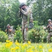 The image shows a group of tactical personnel in military/law enforcement gear moving through an open field with yellow wildflowers and green vegetation. Multiple individuals in camouflage uniforms, tactical vests, and helmets are advancing with weapons raised in a training or operational formation. A person in a dark law enforcement uniform is visible on the left edge, suggesting a joint military-law enforcement exercise.