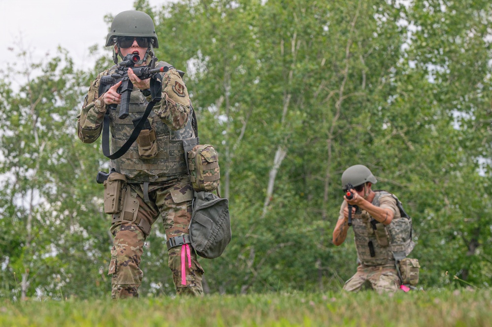Two military personnel in camouflage uniforms are conducting a field training exercise in a wooded outdoor environment. The foreground soldier is standing and aiming a rifle with a red optic attachment, while the background soldier is in a kneeling position also aiming a weapon. Both wear tactical gear including helmets, body armor, and equipment pouches.