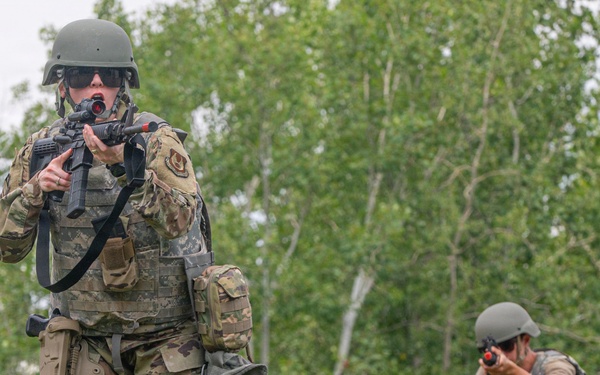 Two military personnel in camouflage uniforms are conducting a field training exercise in a wooded outdoor environment. The foreground soldier is standing and aiming a rifle with a red optic attachment, while the background soldier is in a kneeling position also aiming a weapon. Both wear tactical gear including helmets, body armor, and equipment pouches.