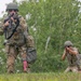 Two military personnel in camouflage uniforms are conducting a field training exercise in a wooded outdoor environment. The foreground soldier is standing and aiming a rifle with a red optic attachment, while the background soldier is in a kneeling position also aiming a weapon. Both wear tactical gear including helmets, body armor, and equipment pouches.