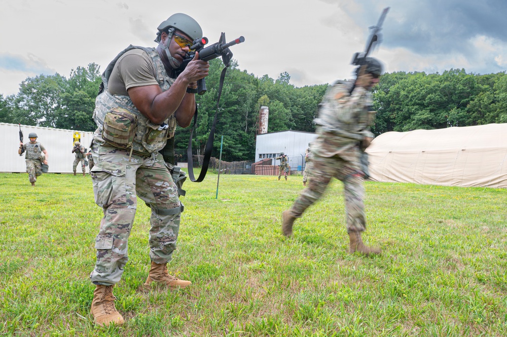 Military personnel in camouflage uniforms are conducting a field training exercise on a grassy area. The foreground soldier is in an aiming stance with a rifle equipped with a red laser/MILES training device, while other soldiers are moving in the background. The setting includes a training facility with tents and modular buildings surrounded by trees.