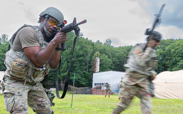 Military personnel in camouflage uniforms are conducting a field training exercise on a grassy area. The foreground soldier is in an aiming stance with a rifle equipped with a red laser/MILES training device, while other soldiers are moving in the background. The setting includes a training facility with tents and modular buildings surrounded by trees.