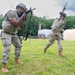 Military personnel in camouflage uniforms are conducting a field training exercise on a grassy area. The foreground soldier is in an aiming stance with a rifle equipped with a red laser/MILES training device, while other soldiers are moving in the background. The setting includes a training facility with tents and modular buildings surrounded by trees.