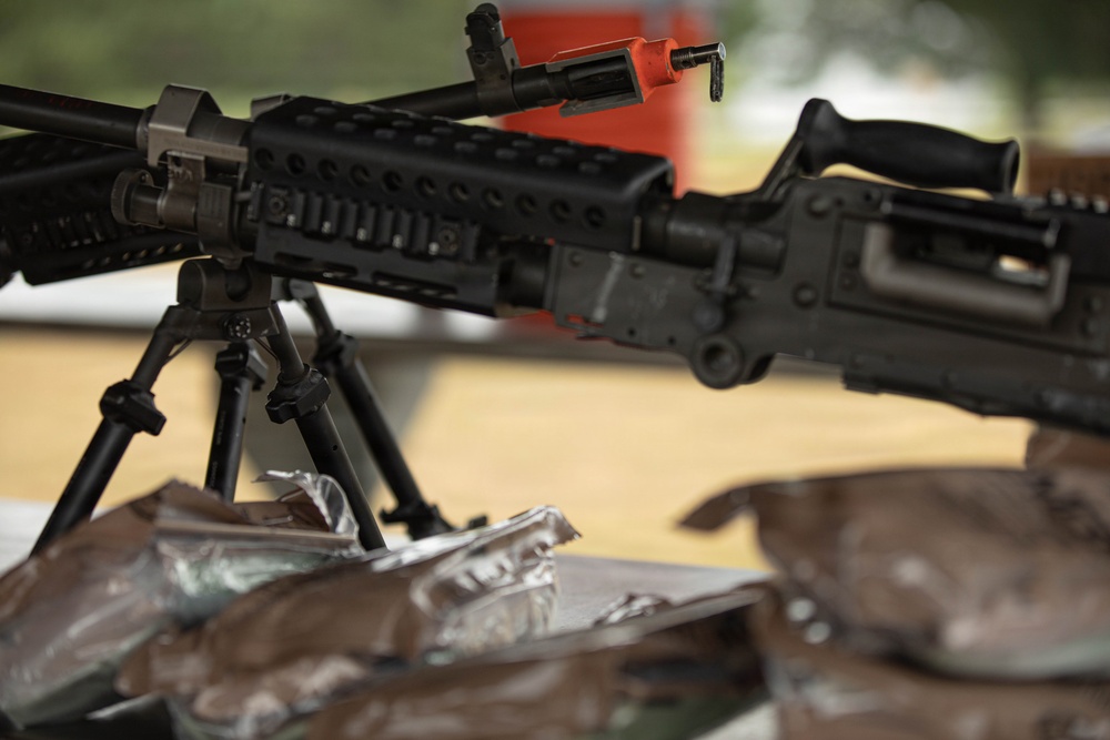 The image shows a military or law enforcement display featuring at least one rifle mounted on a bipod stand on a table. The weapon appears to be an AR/M16-style platform with a Picatinny rail system and a safety/clearing indicator (orange device) attached. In the foreground, there are several dark-colored pouches or packages, possibly military MRE pouches or equipment bags.