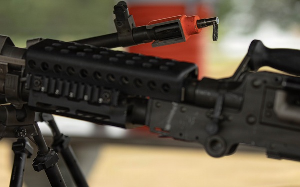 The image shows a military or law enforcement display featuring at least one rifle mounted on a bipod stand on a table. The weapon appears to be an AR/M16-style platform with a Picatinny rail system and a safety/clearing indicator (orange device) attached. In the foreground, there are several dark-colored pouches or packages, possibly military MRE pouches or equipment bags.