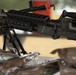 The image shows a military or law enforcement display featuring at least one rifle mounted on a bipod stand on a table. The weapon appears to be an AR/M16-style platform with a Picatinny rail system and a safety/clearing indicator (orange device) attached. In the foreground, there are several dark-colored pouches or packages, possibly military MRE pouches or equipment bags.