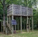 A large elevated wooden platform structure stands in a wooded area, supported by multiple tall wooden posts. The structure features vertical wooden slat walls forming an enclosed upper level, accessible via a wooden staircase on the left side. A solar panel is mounted near the base, suggesting the structure has some electrical power needs.