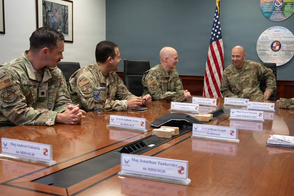 Four U.S. Air Force personnel in OCP (Operational Camouflage Pattern) uniforms are seated around a conference table in what appears to be a military briefing room. Name placards on the table identify several individuals including Lt Col Anthony Aceto (66 MDS/SGN), Lt Col Ryan Gough (66 MDS/SGP), Col Patrick Cannon (Commander, 66 ABG), Colonel Charles Silvanic (Commander, 66 ABG), CMSgt Daniel Anderson (Command Chief, 66 ABG), and MSgt Rodric Peterson (Senior Enlisted Leader). The group appears to be engaged in a relaxed, informal discussion with visible smiling and laughter.