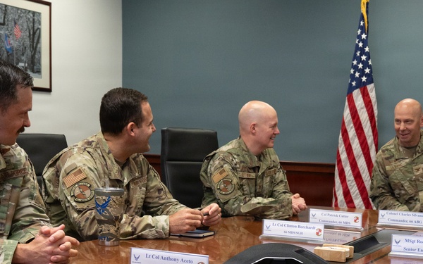 Four U.S. Air Force personnel in OCP (Operational Camouflage Pattern) uniforms are seated around a conference table in what appears to be a military briefing room. Name placards on the table identify several individuals including Lt Col Anthony Aceto (66 MDS/SGN), Lt Col Ryan Gough (66 MDS/SGP), Col Patrick Cannon (Commander, 66 ABG), Colonel Charles Silvanic (Commander, 66 ABG), CMSgt Daniel Anderson (Command Chief, 66 ABG), and MSgt Rodric Peterson (Senior Enlisted Leader). The group appears to be engaged in a relaxed, informal discussion with visible smiling and laughter.