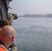 Personnel are leaning out of an open helicopter door observing a body of water below. At least one individual is wearing military camouflage and a flight helmet, while others wear orange safety vests, suggesting a search and rescue or reconnaissance operation. A small boat is visible in the distance on the water, with a treeline obscured by haze in the background.