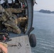 Military personnel in full combat gear are positioned inside the open door of a helicopter hovering low over a body of water. One soldier wearing a flight helmet and multicam uniform is leaning out of the aircraft, appearing to conduct a water-related operation or reconnaissance. The helicopter's landing gear is visible above the water surface, with a forested shoreline visible in the background.