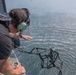 A person wearing military-style communications headset (green aviation headphones) is leaning out from what appears to be a military helicopter or aircraft over open water. They appear to be deploying or releasing a small folded drone or quadcopter frame into the water below. The rotor wash from the aircraft is visibly disturbing the water surface beneath.