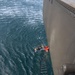 An aerial/overhead view from a large gray military or naval vessel shows two individuals in the water alongside the ship's hull. A rope/Jacob's ladder hangs down the side of the vessel toward the water. One person appears to be wearing a wetsuit and diving mask, while the other is wearing an orange life vest, suggesting a man-overboard rescue drill or actual rescue operation.