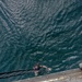 An aerial/overhead photograph taken from the deck or ramp of a military or naval vessel looking down at a swimmer in dark green open water. A person in diving gear including a mask and wetsuit is swimming toward a rope ladder hanging from the vessel. A second person's hand is visible in the foreground gripping the edge of the metal ramp or deck.