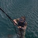 An aerial photograph taken from above shows a water rescue or training operation in open water. A person in military/tactical gear, wearing diving goggles and a life vest, is climbing a rope ladder suspended from above while two individuals in orange inflatable life vests float in the water below. The scene appears to be a military or search-and-rescue training exercise conducted from a vessel or aircraft.