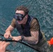 A man wearing a diving mask and tactical vest is being pulled out of open water, gripping a rope or handle extended from above. He has extensive tattoos on his right arm and is wearing a watch and olive-colored shirt. A second person in camouflage and an orange life vest is partially visible in the lower right corner.