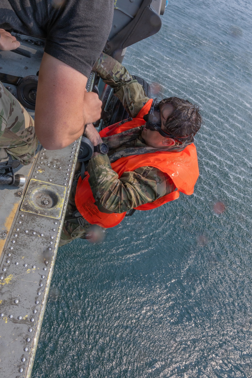 A military or rescue personnel wearing camouflage uniform and an orange inflatable life vest is being hoisted or assisted at the edge of what appears to be a helicopter or aircraft over open water. Another person in camouflage uniform is gripping the individual, likely assisting in a hoist rescue or water survival training exercise. The subject appears wet, suggesting recent water immersion.