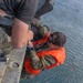 A military or rescue personnel wearing camouflage uniform and an orange inflatable life vest is being hoisted or assisted at the edge of what appears to be a helicopter or aircraft over open water. Another person in camouflage uniform is gripping the individual, likely assisting in a hoist rescue or water survival training exercise. The subject appears wet, suggesting recent water immersion.