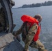 A military personnel wearing camouflage uniform and an inflated orange life vest is leaning out from the open door of a military helicopter over a body of water. A second crew member in full flight gear and helmet is visible inside the aircraft. The scene appears to depict a water rescue or helicopter water operations training exercise.