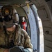 A U.S. Army soldier is seated inside a military helicopter, wearing a flight helmet with a 'Charlie Don't Surf' patch and integrated communications equipment. The soldier is dressed in OCP (Operational Camouflage Pattern) uniform with a U.S. flag patch visible on the left shoulder. The image captures the interior of the aircraft with safety equipment visible in the background.