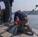 A military helicopter crew member in multicam/OCP camouflage uniform is kneeling at the open rear ramp of a helicopter, assisting a person wearing a red life vest or rescue garment. The scene appears to be a water rescue or training operation over a body of water, with a small patrol boat visible in the background. A second crew member is seated inside the helicopter cabin to the left.