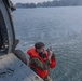 A military or rescue operation is underway involving a helicopter hovering low over a body of water. A person in camouflage uniform and an orange life vest is positioned at the open door of the helicopter, appearing to signal with their hand. A second person in full tactical gear is seated inside the helicopter observing the operation.
