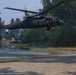 A U.S. Army UH-60 Black Hawk helicopter is flying at low altitude over a shoreline area near a body of water. The helicopter has its side door open with at least one crew member visible in tactical gear. The rotor wash is visibly disturbing the water surface below the aircraft.