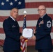 Two U.S. Air Force personnel are participating in a formal military ceremony, jointly holding a certificate that appears to be a Certificate of Retirement. The ceremony is taking place in what appears to be a hangar or large indoor venue decorated with a large American flag as a backdrop. The individual on the right, wearing a Chief Master Sergeant rank insignia and multiple service ribbons with a medal, appears to be the retiree receiving the certificate.