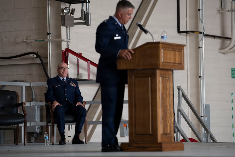 A military ceremony is taking place inside what appears to be an aircraft hangar or military facility. One uniformed Air Force officer stands at a wooden podium delivering a speech, while a senior non-commissioned officer sits in a chair on the stage behind him. The setting and formal dress uniforms suggest this is an official military ceremony such as a promotion, retirement, or change of command.