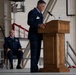 A military ceremony is taking place inside what appears to be an aircraft hangar or military facility. One uniformed Air Force officer stands at a wooden podium delivering a speech, while a senior non-commissioned officer sits in a chair on the stage behind him. The setting and formal dress uniforms suggest this is an official military ceremony such as a promotion, retirement, or change of command.