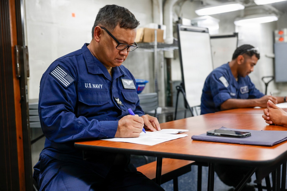 Two U.S. Navy personnel in blue coverall uniforms are seated at wooden tables in what appears to be an interior compartment of a naval vessel. The foreground sailor, wearing glasses, is actively writing on documents with a blue pen. A second sailor is visible in the background, also seated at a table.