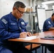 Two U.S. Navy personnel in blue coverall uniforms are seated at wooden tables in what appears to be an interior compartment of a naval vessel. The foreground sailor, wearing glasses, is actively writing on documents with a blue pen. A second sailor is visible in the background, also seated at a table.