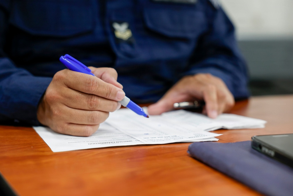 A uniformed individual, likely a military or law enforcement officer based on visible insignia and dark blue uniform, is writing on official documents at a wooden desk. The person holds a blue pen and appears to be filling out or reviewing paperwork. Additional items including a dark device and a blue folder are visible on the desk surface.