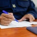 A uniformed individual, likely a military or law enforcement officer based on visible insignia and dark blue uniform, is writing on official documents at a wooden desk. The person holds a blue pen and appears to be filling out or reviewing paperwork. Additional items including a dark device and a blue folder are visible on the desk surface.