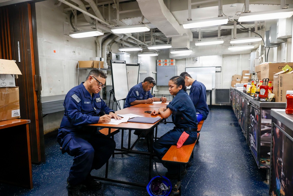 Four U.S. Navy personnel in dark blue uniforms are seated at tables in what appears to be a mess hall or galley aboard a naval vessel. They appear to be engaged in paperwork or a briefing session. The setting is characterized by industrial overhead piping, fluorescent lighting, and condiment supplies visible along a counter on the right.