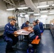 Four U.S. Navy personnel in dark blue uniforms are seated at tables in what appears to be a mess hall or galley aboard a naval vessel. They appear to be engaged in paperwork or a briefing session. The setting is characterized by industrial overhead piping, fluorescent lighting, and condiment supplies visible along a counter on the right.
