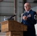 A senior U.S. Air Force enlisted member in dress blues is delivering a speech at a podium inside what appears to be a military hangar. The individual displays numerous service ribbons, medals, and senior enlisted rank insignia consistent with a Chief Master Sergeant. The setting suggests a formal military ceremony, likely a retirement or change of command event.