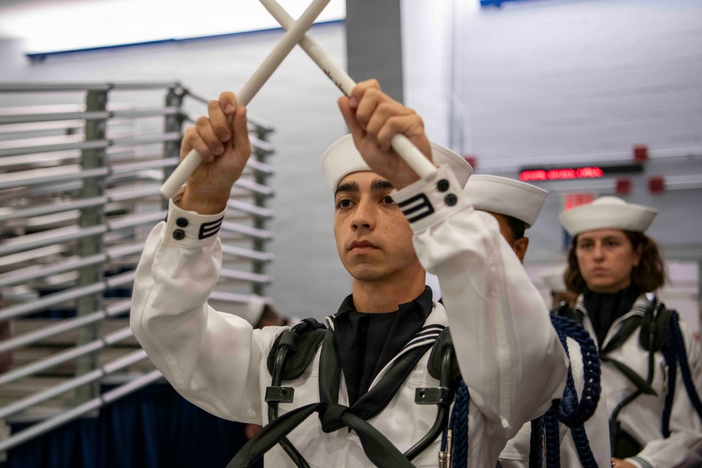 A young person in a white naval/military-style uniform is performing a drum major or color guard routine, holding two white batons crossed above their head. Additional uniformed individuals are visible in the background. The setting appears to be an indoor facility, possibly a gymnasium or convention hall.