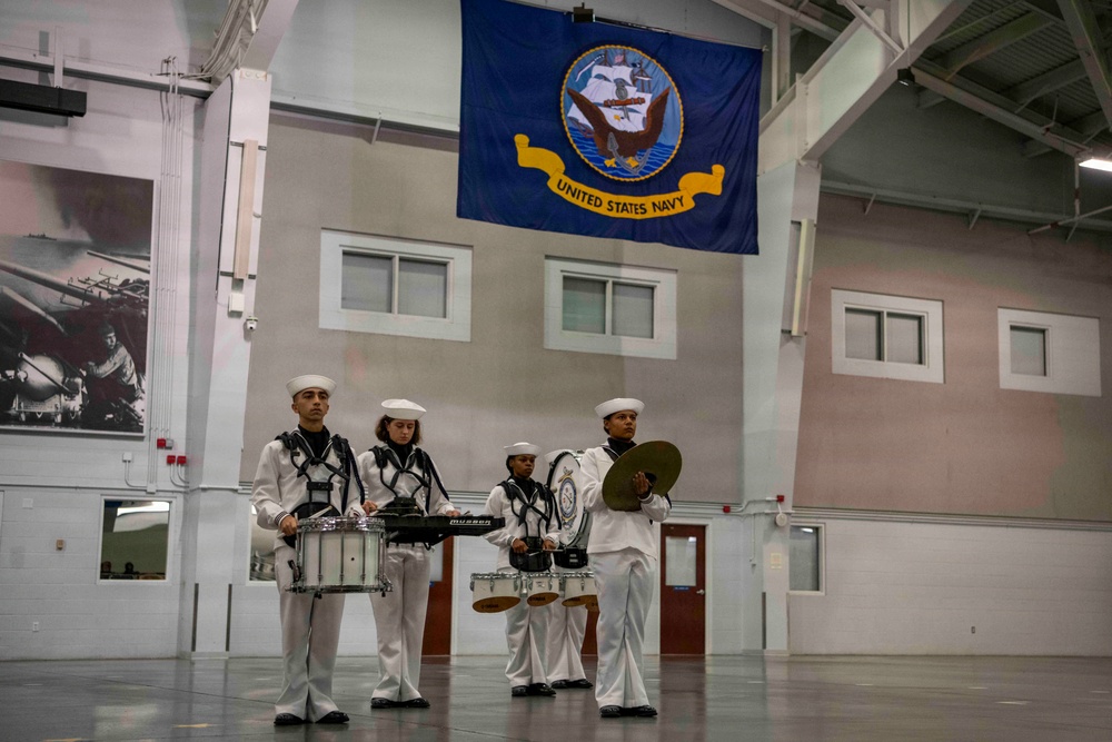 Four United States Navy personnel in white dress uniforms perform as a percussion ensemble inside a large indoor facility. They are playing snare drums and cymbals beneath a prominently displayed United States Navy flag. The setting appears to be a military hangar or drill hall with historical imagery visible on the left wall.