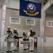 Four United States Navy personnel in white dress uniforms perform as a percussion ensemble inside a large indoor facility. They are playing snare drums and cymbals beneath a prominently displayed United States Navy flag. The setting appears to be a military hangar or drill hall with historical imagery visible on the left wall.