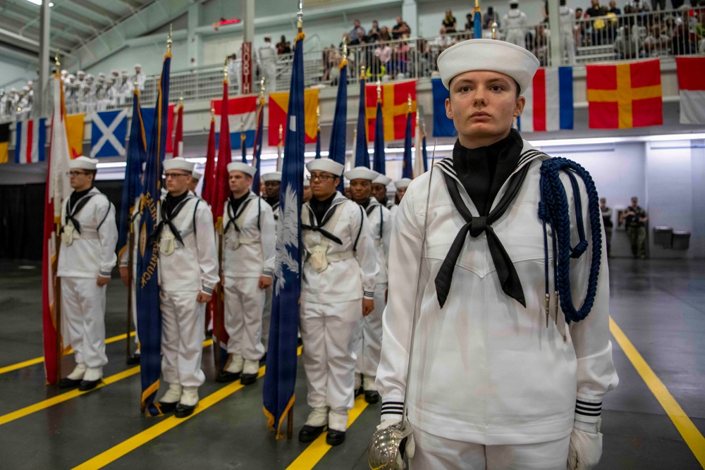 A formal U.S. Navy military ceremony, likely a graduation or pass-in-review, taking place inside a large indoor facility. Sailors in dress white uniforms are standing at attention in formation, with a color guard holding multiple state and military flags. Spectators are visible in elevated bleachers in the background.