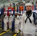 A formal U.S. Navy military ceremony, likely a graduation or pass-in-review, taking place inside a large indoor facility. Sailors in dress white uniforms are standing at attention in formation, with a color guard holding multiple state and military flags. Spectators are visible in elevated bleachers in the background.