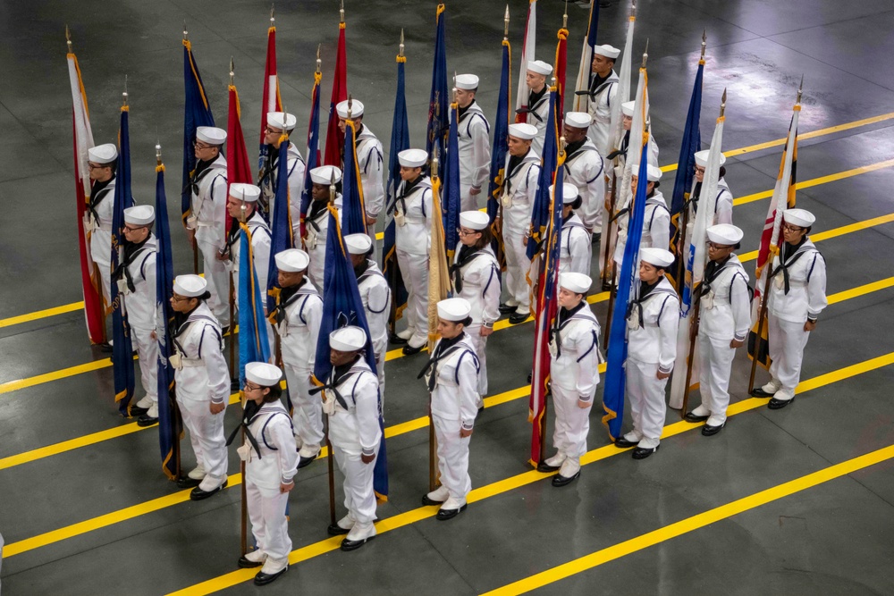 A large formation of U.S. Navy sailors in dress white uniforms are standing at attention in an indoor drill hall or gymnasium, each carrying a ceremonial flag on a pole. The formation appears to be part of a formal military ceremony, likely a graduation or pass-in-review event. Yellow lane markings are visible on the gray concrete floor, suggesting a large multipurpose facility.