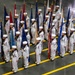 A large formation of U.S. Navy sailors in dress white uniforms are standing at attention in an indoor drill hall or gymnasium, each carrying a ceremonial flag on a pole. The formation appears to be part of a formal military ceremony, likely a graduation or pass-in-review event. Yellow lane markings are visible on the gray concrete floor, suggesting a large multipurpose facility.