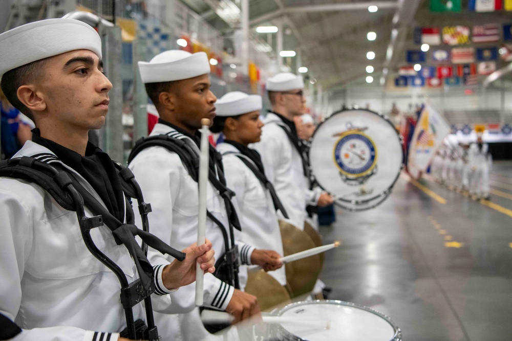 A military marching band, dressed in white U.S. Navy dress uniforms, is performing at an indoor ceremony or graduation event. The drummers are lined up in formation, holding drumsticks and playing snare drums, with a large bass drum bearing a naval insignia visible in the background. The setting appears to be a large indoor facility decorated with state and military flags.