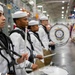 A military marching band, dressed in white U.S. Navy dress uniforms, is performing at an indoor ceremony or graduation event. The drummers are lined up in formation, holding drumsticks and playing snare drums, with a large bass drum bearing a naval insignia visible in the background. The setting appears to be a large indoor facility decorated with state and military flags.